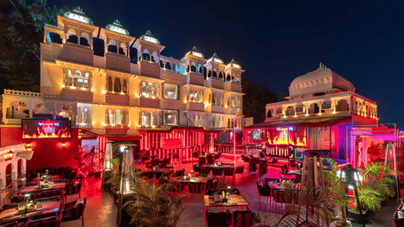 Night-time view of a white hotel exterior, showcasing an outdoor dining area with red-themed lighting and seating.