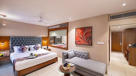A spacious hotel room  featuring a large double bed with decorative pillows, a grey chaise lounge, and modern artwork on the wall -  SK Lords Inn Ahmedabad