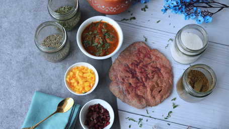 A flatbread served with curry, diced vegetables, and jars of assorted spices on a wooden table.