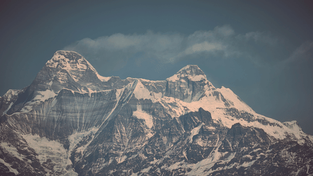 Snow-dusted mountain peaks rise dramatically beneath a moody, atmospheric sky filled with soft clouds.