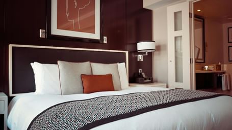 Elegantly decorated hotel bedroom with a large bed, multiple pillows, a patterned bed runner, artwork above the headboard, and a glimpse into an adjoining room.