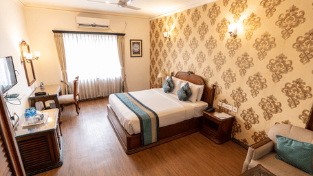 A hotel room setup featuring a king size bed, a wardrobe, a work desk with a chair, a television unit, and a ceiling fan at The Lindsay, Kolkata.