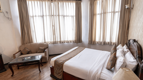 A hotel room with a king size bed, large curtained windows, and a sofa with a coffee table at The Lindsay, Kolkata.