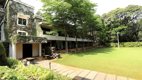 Exterior of a 2-storey stone building at Trance Sunderban Resort surrounded by tall green trees and a sunlit paved walkway.