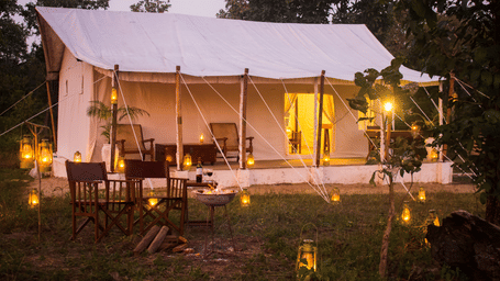 Exterior view of a traditional safari tent illuminated from within at dusk, with warm string lights and folding chairs out front - Umaria Safari Camp