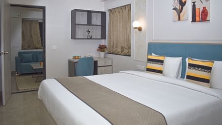 A hotel bedroom with a bed, a desk and chair, a wall cabinet, a window with curtains, and a doorway leading to a living area with a blue sofa - Urban Suites by BluSalzz, Jaipur