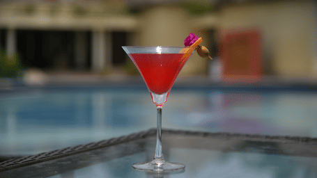 A close-up of a red martini cocktail glass placed on the edge of the swimming pool at Vedic Village Spa Resort with a blurred pool background in a warm outdoor setting.