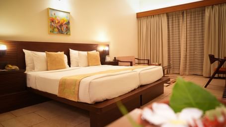 Neatly arranged double bed in the Standard Suite at Vedic Village Spa Resort with a wooden headrest, a painting above the bed, and large window drapes drawn shut.