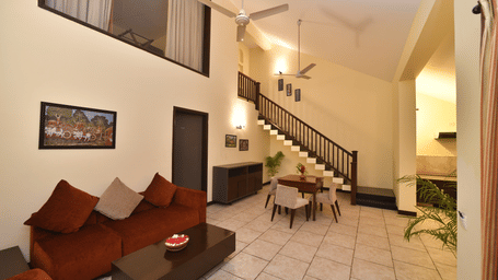 A wide angle view of a villa interior at Vedic Village Spa Resort featuring a wooden staircase cushioned brown sofas tiled flooring ceiling fan and warm ambient lighting throughout.