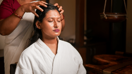 Therapist performing a seated ayurvedic scalp massage on a female guest in a white robe at Vedic Village Spa Resort Kolkata with warm ambient lighting.