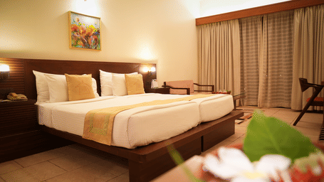 Neatly arranged double bed in the Standard Suite at Vedic Village Spa Resort with a wooden headrest, a painting above the bed, and large window drapes drawn shut.