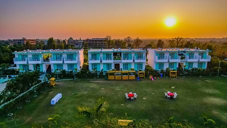 Sunset view of Vedikant Resorts The Mallard Corbett with the building facing a lawn setup for outdoor dining and the sun near the horizon.