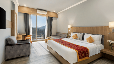 A hotel room with a bed, seating area, desk, and view through a large window - Via Lakhela Resort & Spa, Kumbhalgarh