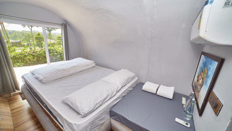 The interior of a modern, capsule-style accommodation unit featuring a single bed with a white mattress and a small bedside table, with a window offering a view of greenery. - Vintage Trails by Ilara