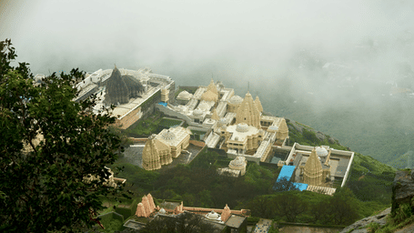 Mist-covered hillside surrounding Ambaji Temple complex, with white buildings emerging through clouds and dense greenery during early morning light
