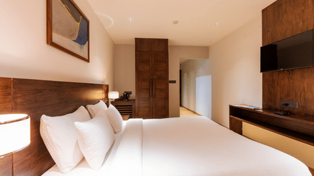 A side view of Superior Room of Visthara with a double bed, a flat-screen TV mounted to wall, modern wooden furnishing, and a side lamp glowing