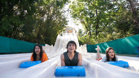 3 people enjoying What-A-Coaster ride at Water Kingdom while sliding down with a mat and trees on either side of the ride.