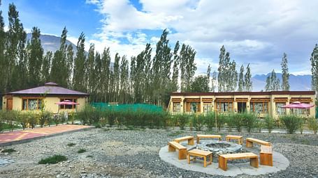 An overview of White Sands Resort, Ladakh featuring a communal seating area with wooden benches and a fire pit surrounded by gravel, with cottages and trees in the background under a cloudy sky.
