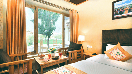 Standard Cottage with a king size bed, a window with orange curtains, a small table and chairs, and a painting on the wall at White Sands Resort, Ladakh.