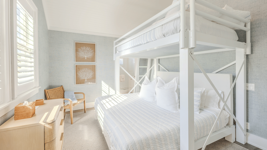 A wide view of a light-filled bedroom featuring a large white bunk bed, a light wood dresser, and minimalist coastal decor.