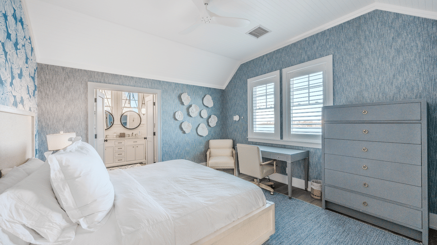 A bedroom with blue patterned wallpaper, a gray dresser, and a desk area, with a doorway leading to a private bathroom.