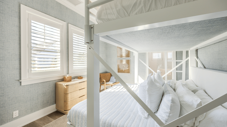 A close-up of a white wooden bunk bed with crisp white linens in a room with light blue walls and plantation shutters.
