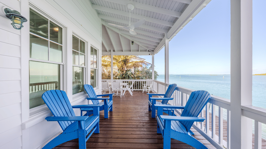A spacious white covered porch with dark wood decking, featuring vibrant blue Adirondack chairs positioned to look out over the clear blue ocean.