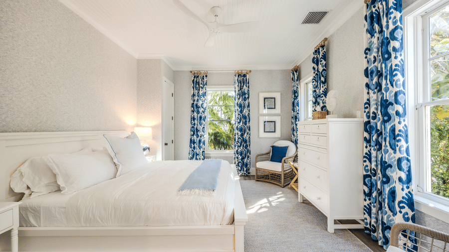 A bright bedroom with all-white bedding, a white dresser, and tall windows framed by bold blue and white patterned curtains