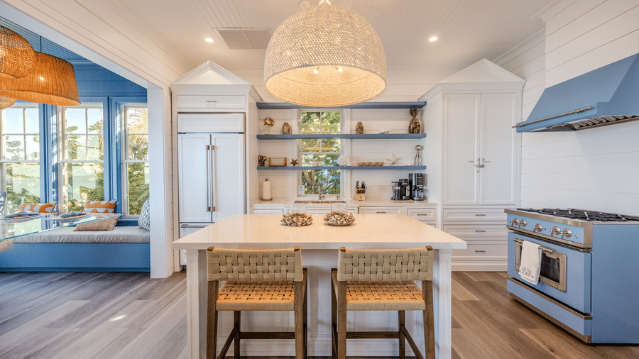 A luxury kitchen featuring a large white island with woven barstools, a professional-grade blue gas range, and open shelving.