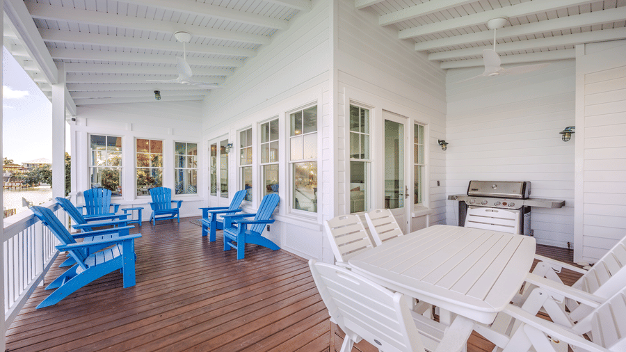 A wide-angle view of a white wrap-around porch featuring a white outdoor dining set, a grill, and blue Adirondack chairs against a backdrop of white siding and large windows.