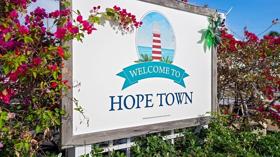 A 'Welcome to Hope Town' sign with a red-and-white striped lighthouse illustration, surrounded by green foliage and pink flowers | Abaco Inn