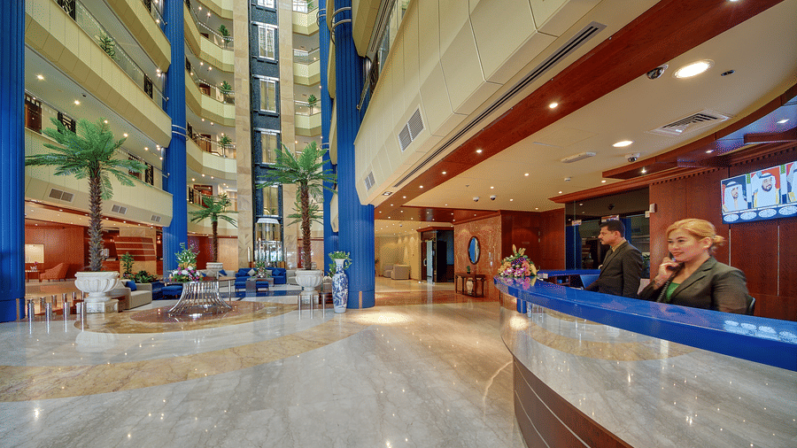 The hotel lobby features a curved reception desk, indoor palm trees, and a multi-storey atrium with blue columns - Al Manar Grand Hotel Apartment, Bur Dubai