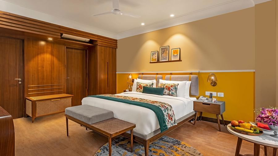 An image of a room featuring a king-size bed with a headboard and comfortable bedding. A bench sits at the foot of the bed, and a small table with a fruit platter is placed nearby.