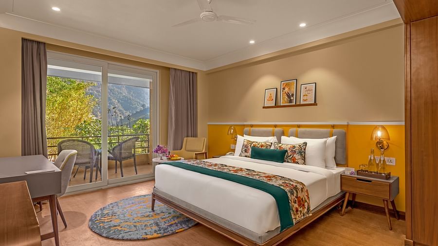 Image of a  room features a large king-size bed with a plush headboard and comfortable bedding. A comfortable armchair sits by a large window offering breathtaking views. The room also includes a wooden desk with a chair, a television, and a ceiling fan. T
