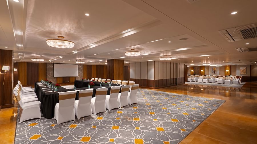 This image shows a large, elegant conference room or banquet hall. The room features high ceilings, a chandelier, and a plush carpeted floor. A long rectangular table is set up in the center of the room,