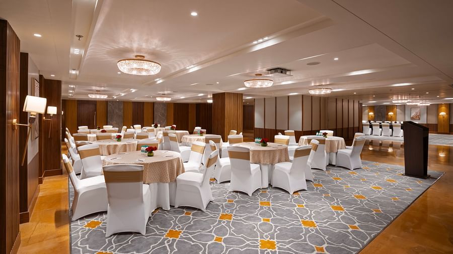 A spacious banquet hall with round tables covered in white tablecloths and chairs. A podium in the background, and the room is decorated with chandeliers and wall lamps.
