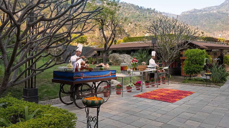 Two chefs are preparing food at outdoor food stations. There are mountains in the background and lush surrounding the scene.