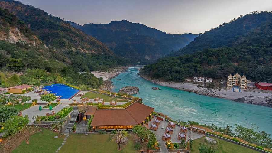 A panoramic view of Aloha on the Ganges, Rishikesh. The image shows the resort nestled amidst hills, with the Ganges River flowing peacefully in the foreground. There is a swimming pool, restaurant, and other facilities visible.