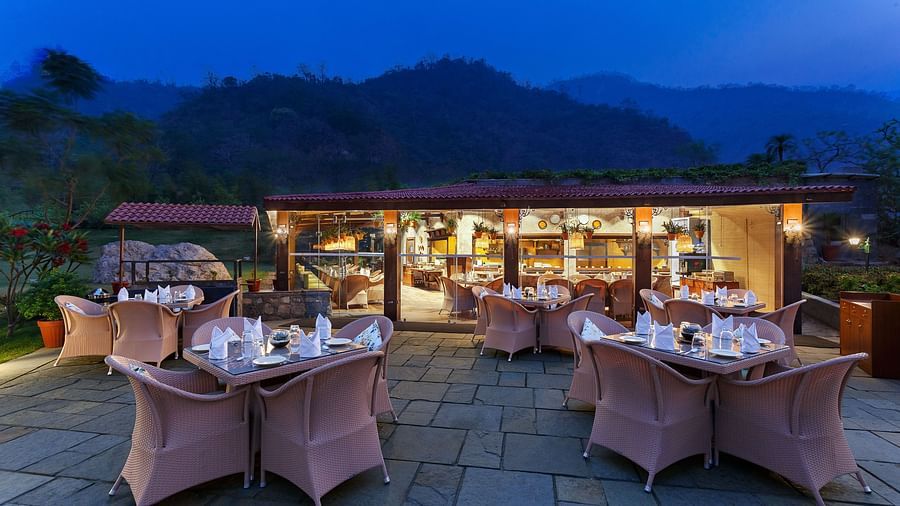 An outdoor restaurant with a view of mountains at dusk. Tables and chairs are set up, a covered area with warm lighting in the background.