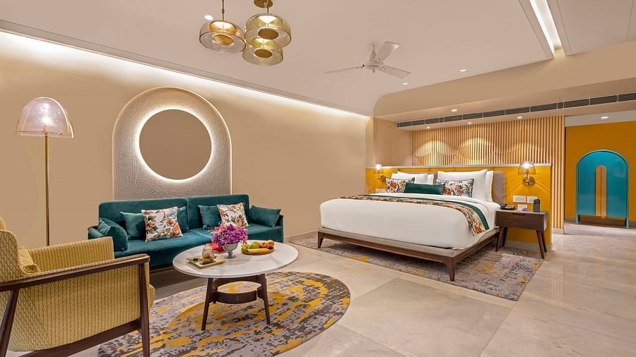 An image of a room featuring a king-size bed, a comfortable sofa, a coffee table, and a television. The room is decorated with a circular mirror, a ceiling fan, and stylish lighting fixtures. 