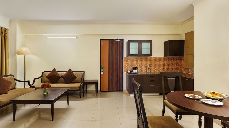 An image of a spacious and modern hotel suite. The living area features a comfortable sofa with throw pillows, a coffee table, and a floor lamp. A separate dining area