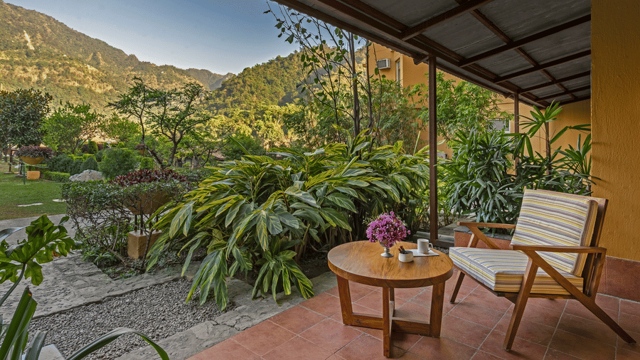 Serene patio at Aloha on the Ganges Resort with wooden chair, table with flowers, tiled floor, lush greenery, and mountain view under clear blue sky.