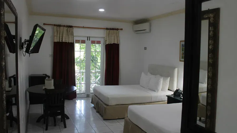 A bright hotel room displays a large bed with white linens, a dark dresser, and a seating area with a table and chairs | Deluxe Double Room | Altamont Court, Jamaica