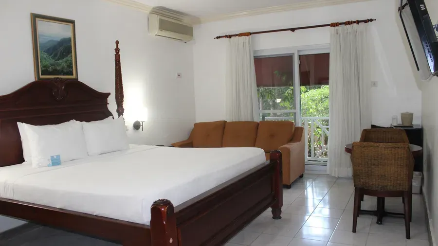 A hotel room features a large wooden bed with white linens, a dark dresser with a television, and a seating area with a couch | Deluxe King Room | Altamont Court, Jamaica