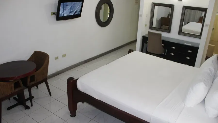 A hotel room presents a bed with white linens, a dark dresser with framed pictures, and a seating area with a table and chairs | Deluxe King Room | Altamont Court, Jamaica