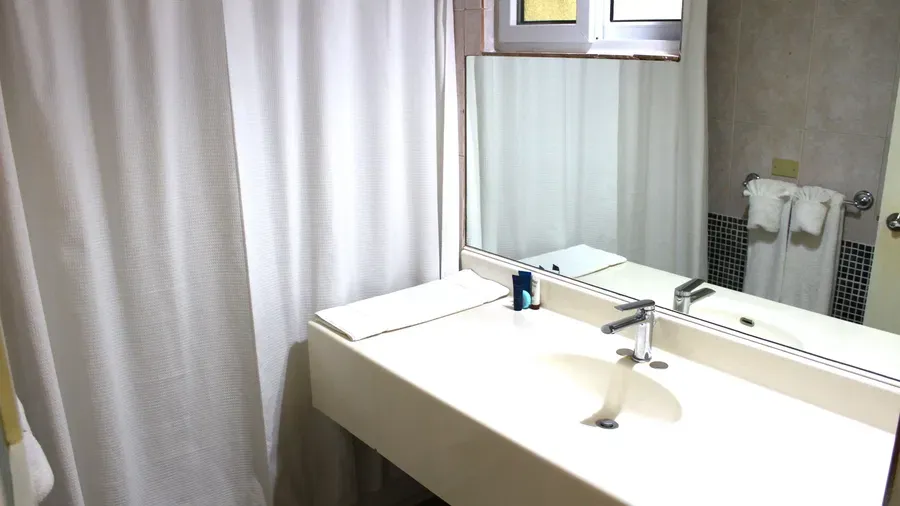 A clean, minimalist bathroom includes a sink with a large mirror and white shower curtains | Executive Double Room | Altamont Court, Jamaica