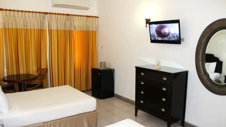 A hotel room showcases a bed with a white comforter, a dark dresser with a television, and yellow curtains by a window | Deluxe Double Room | Altamont Court, Jamaica