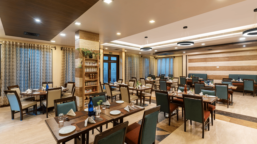 A spacious dining room at Navidya Restaurant featuring modern wooden furniture, polished floors, and large windows that fill the space with light - Amantra Comfort Hotel
