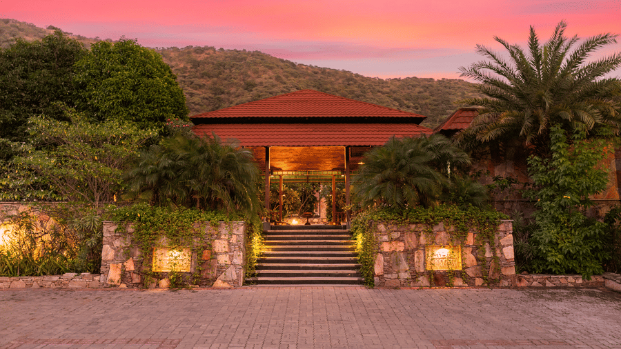 The resort entrance with red roofs and stone walls illuminated against a pink twilight sky | Anandam Jungle Retreat Resort, Udaipur