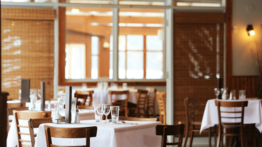 The bright interior of a restaurant with wooden tables and chairs, crisp white tablecloths, and bamboo window blinds.